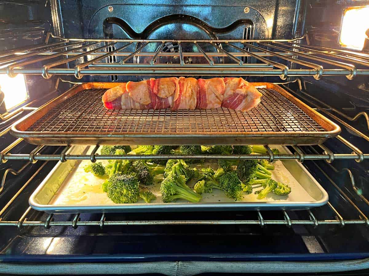 Bacon-wrapped pork tenderloin and broccoli are being baked together in the same oven.