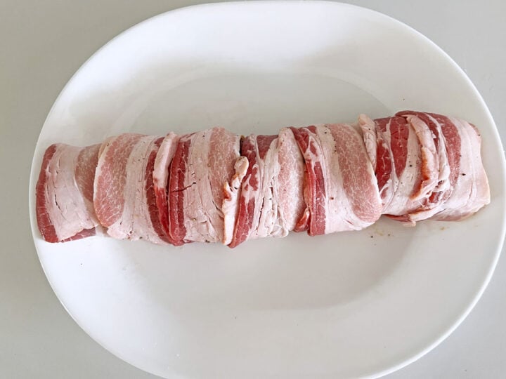 The tenderloin is on a white plate, after it was wrapped in bacon.