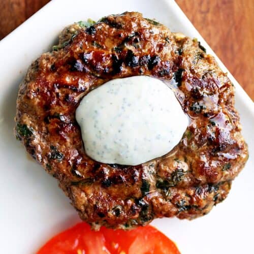 A lamb burger is topped with a yogurt sauce and served with tomato slices.