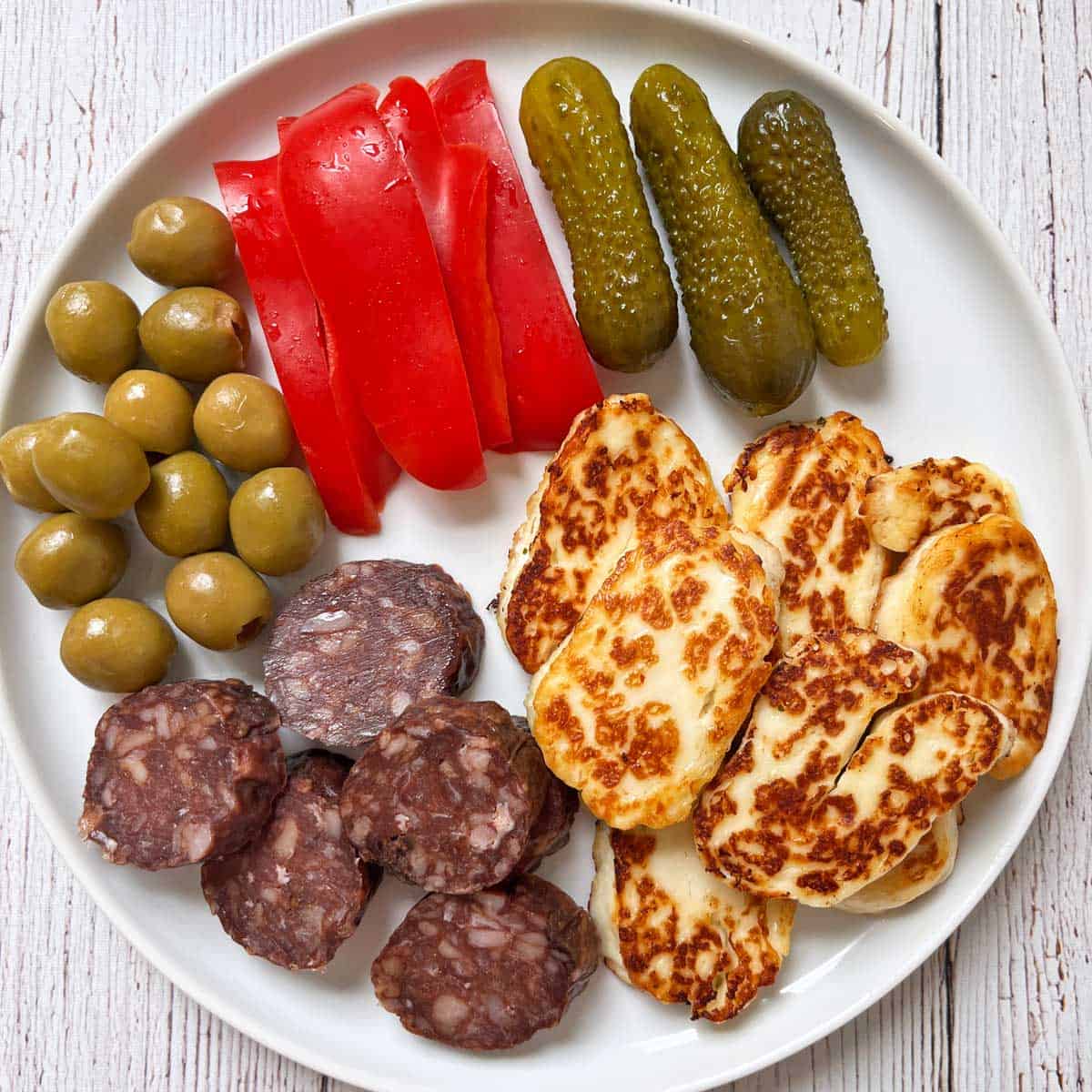 A snack plate with halloumi cheese, salami, peppers, and pickles.