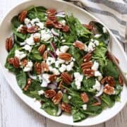 Spinach salad is served on a white platter with a serving spoon.