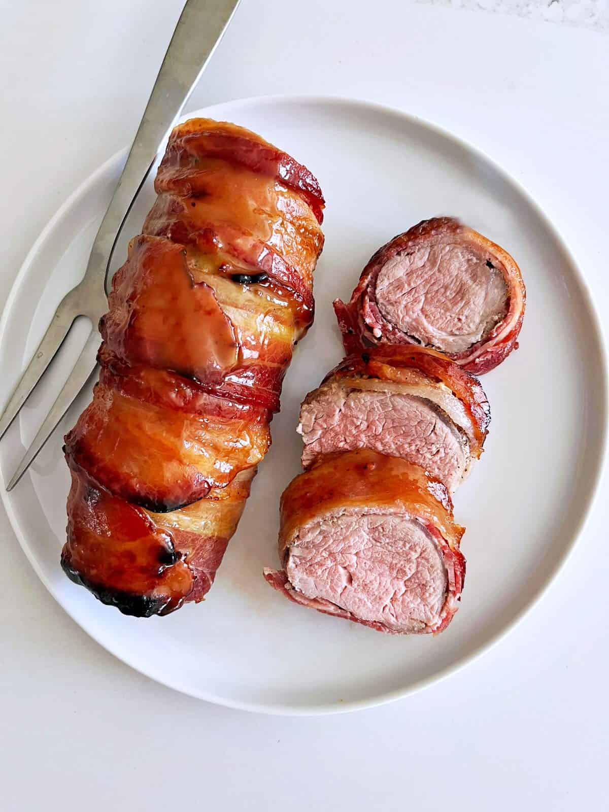 Bacon wrapped pork tenderloin is served on a white plate.
