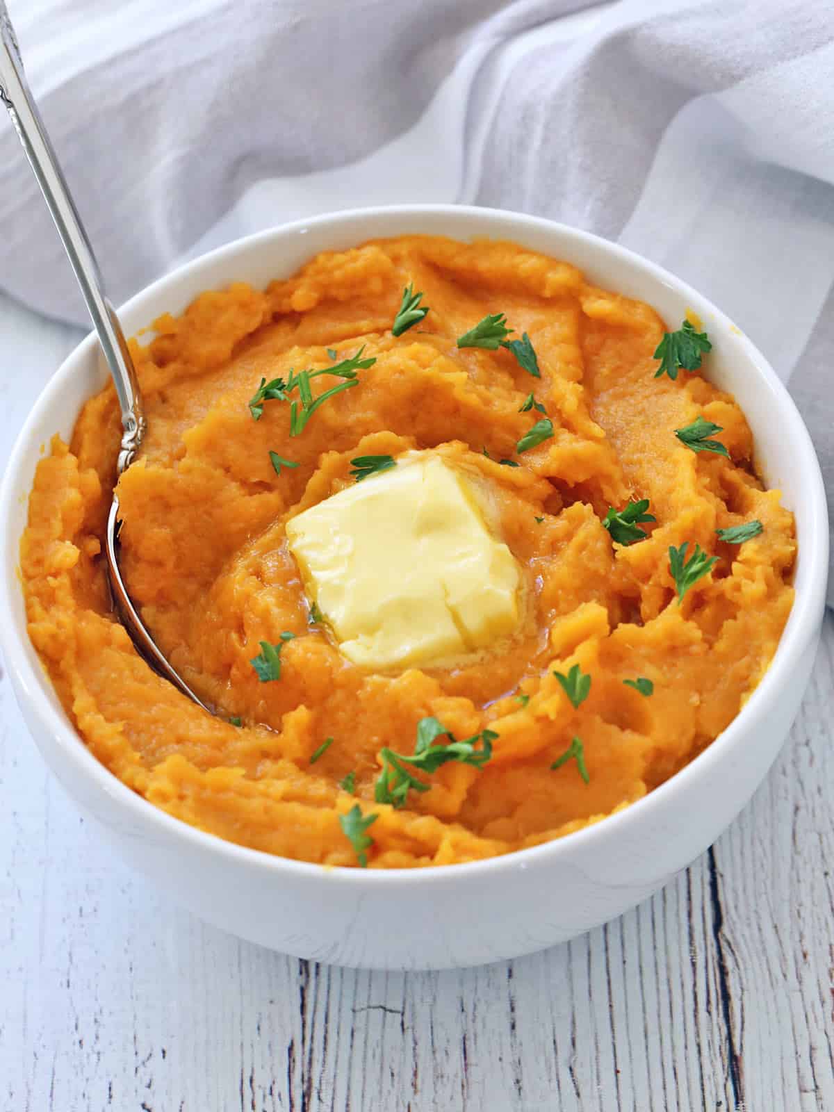 Mashed sweet potatoes are served in a white bowl, topped with butter and garnished with chopped parsley.