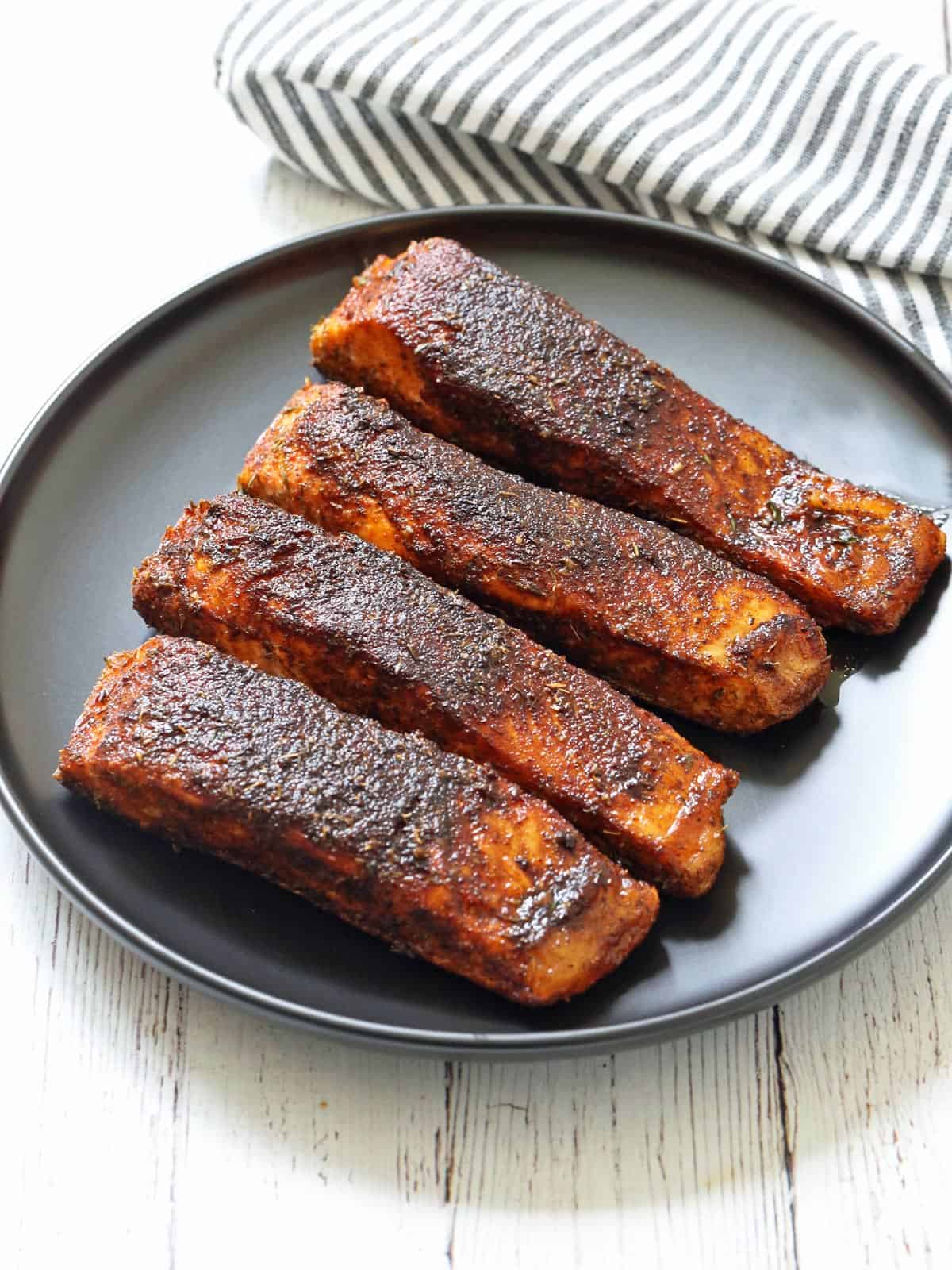 Four pieces of blackened salmon are served on a black plate.