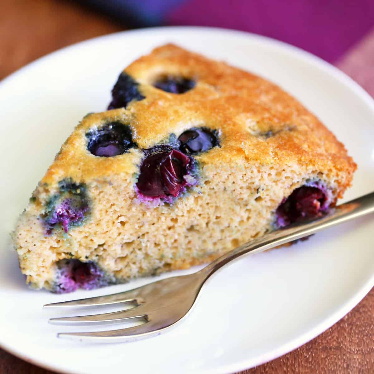 A slice of almond flour blueberry cake is served on a plate with a fork.