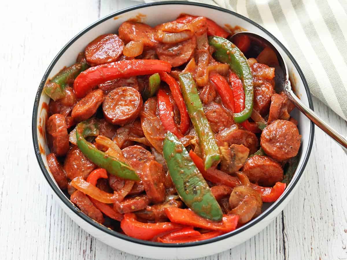 A saucy version of sausage and peppers is served in a white bowl with a serving spoon.