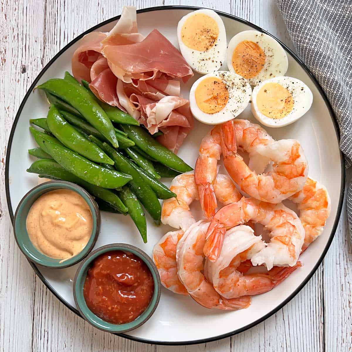 A platter with boiled shrimp, cocktail sauce, sriracha mayo, sugar snap peas, and prosciutto.
