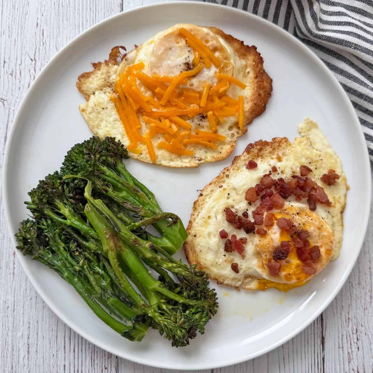 Fried eggs are topped with bacon and cheese and served with broccolini.