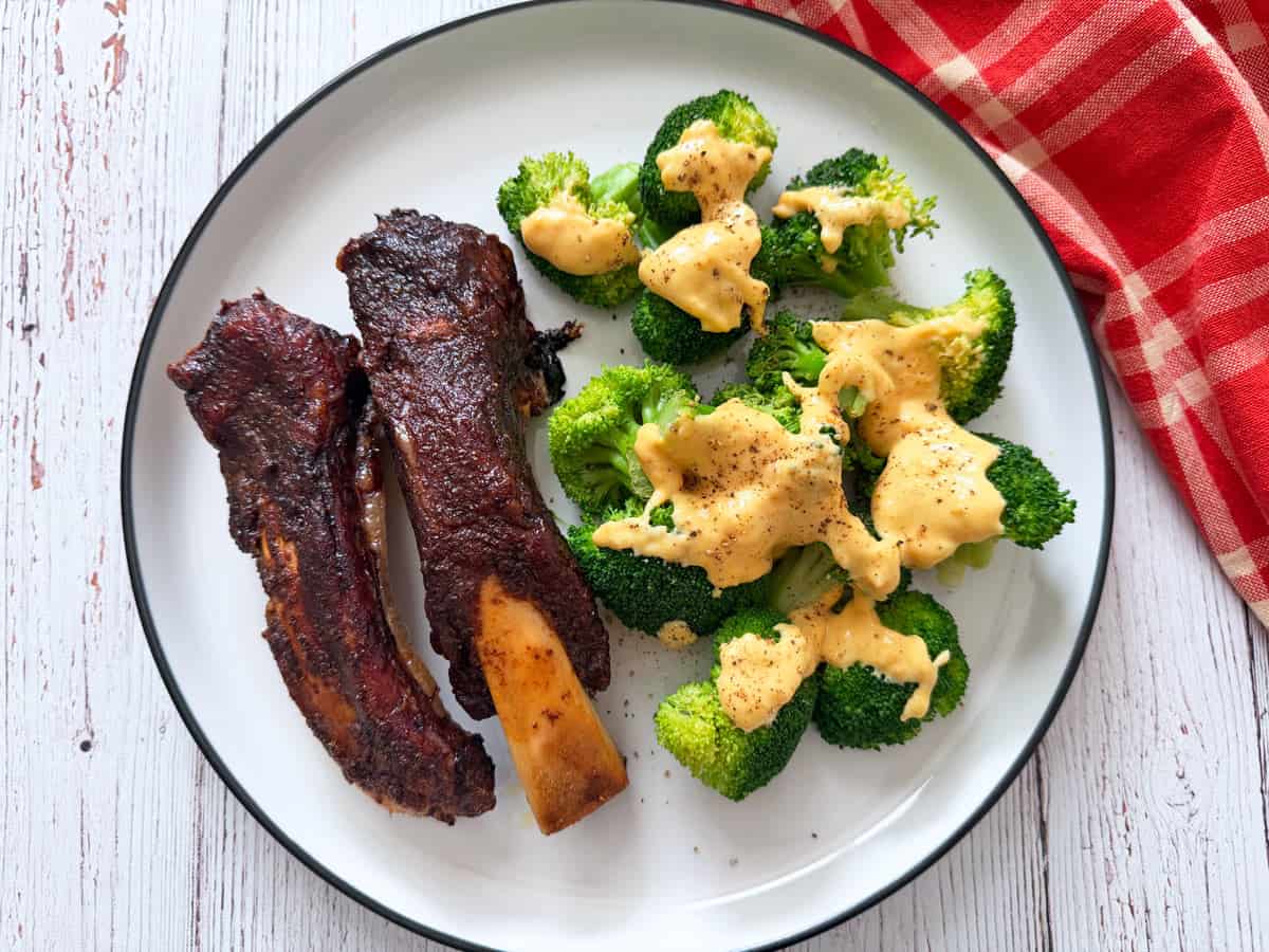 Two beef back ribs are served on a plate with a side of broccoli and cheese.