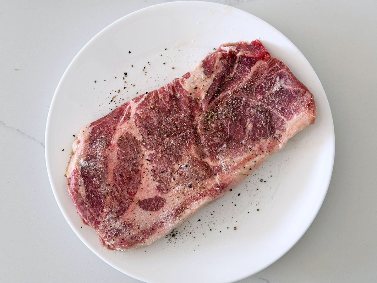 A pork steak is seasoned with coarse salt and black pepper.