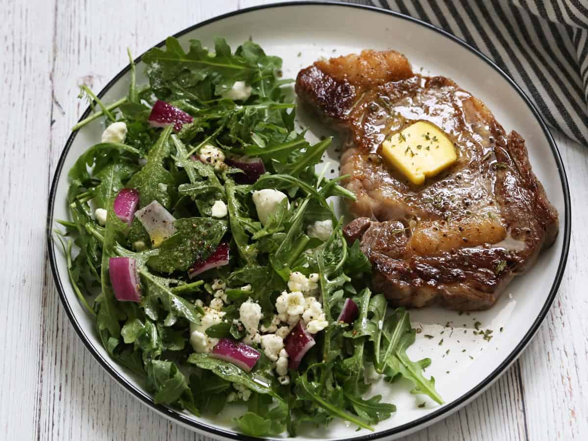 A ribeye steak is served with arugula salad.