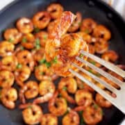 Sauteed shrimp are served in a skillet, and a fork is lifting up one of them.