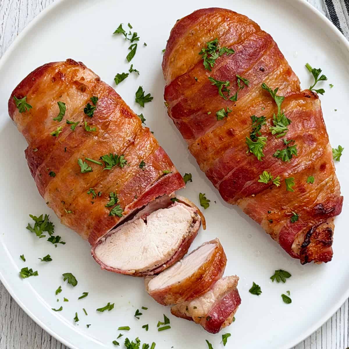 Bacon wrapped chicken breast is served on a plate, topped with parsley.