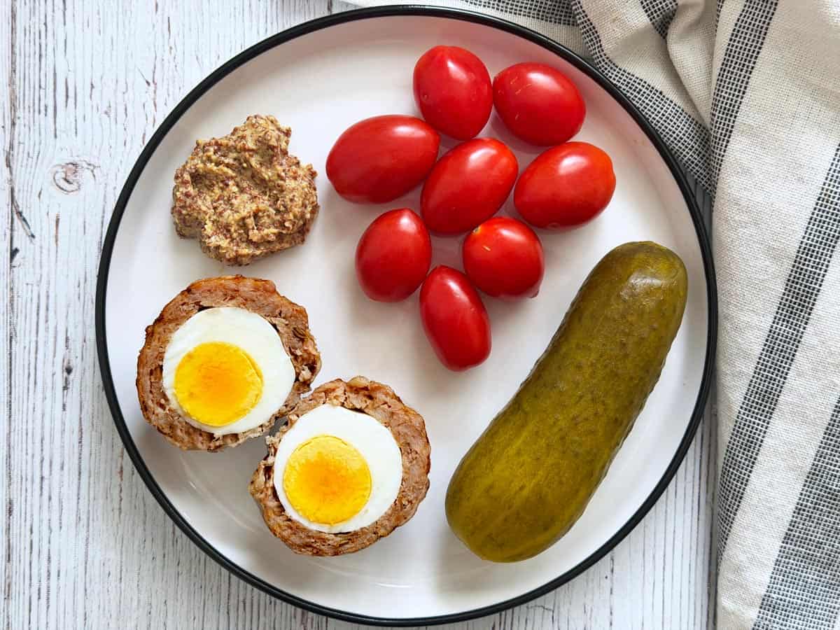 A cold Scotch egg is served with tomatoes, a dill pickle, and grainy mustard.