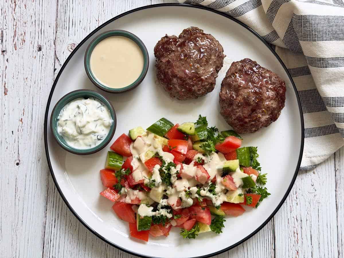 Tzatziki is served on a plate with lamb burgers and a salad.